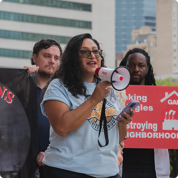 A woman speaking into a megaphone during an outdoor rally, with two people standing behind her holding signs about housing and neighborhood issues. Urban buildings are visible in the background.