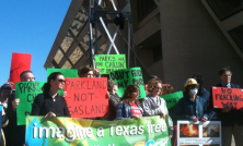 A group of demonstrators gathered outside a large building, holding colorful signs opposing gas drilling and fracking. Messages include “PARKLAND NOT GASLAND,” “PARKS ARE FOR CHILLIN’ NOT DRILLIN’,” and “NO FRACKING WAY.” A large banner in front reads “imagine a Texas free…” as part of the protest display.